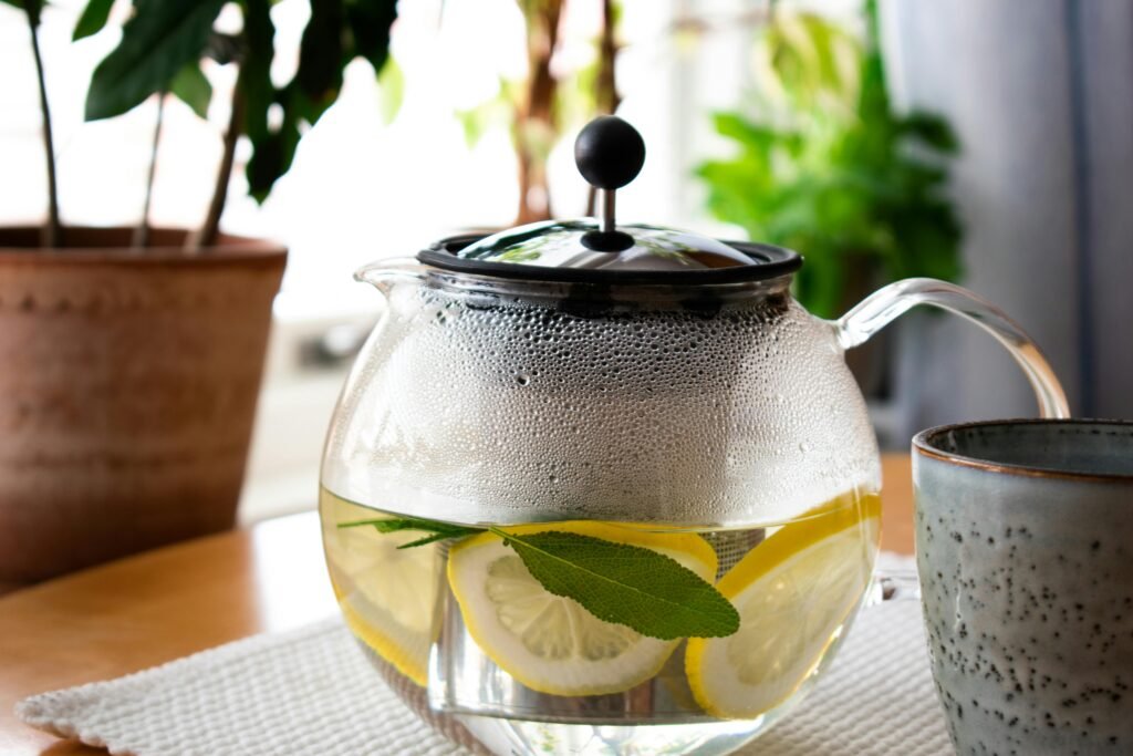 Steaming glass teapot with lemon slices and fresh herbs on a cozy indoor table setting.