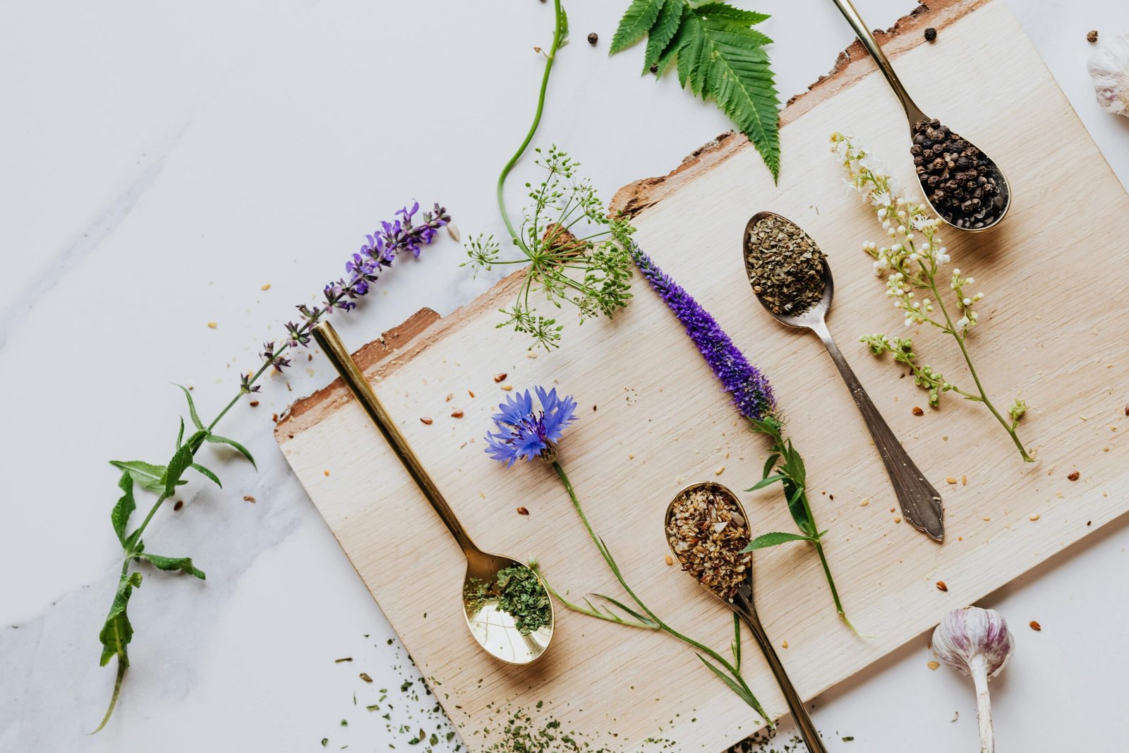 An artistic flat lay of herbs and flowers with teaspoons on a wooden board, perfect for culinary and health themes.